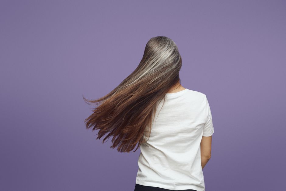 Rear view of a woman with long, flowing gray hair against a purple backdrop.